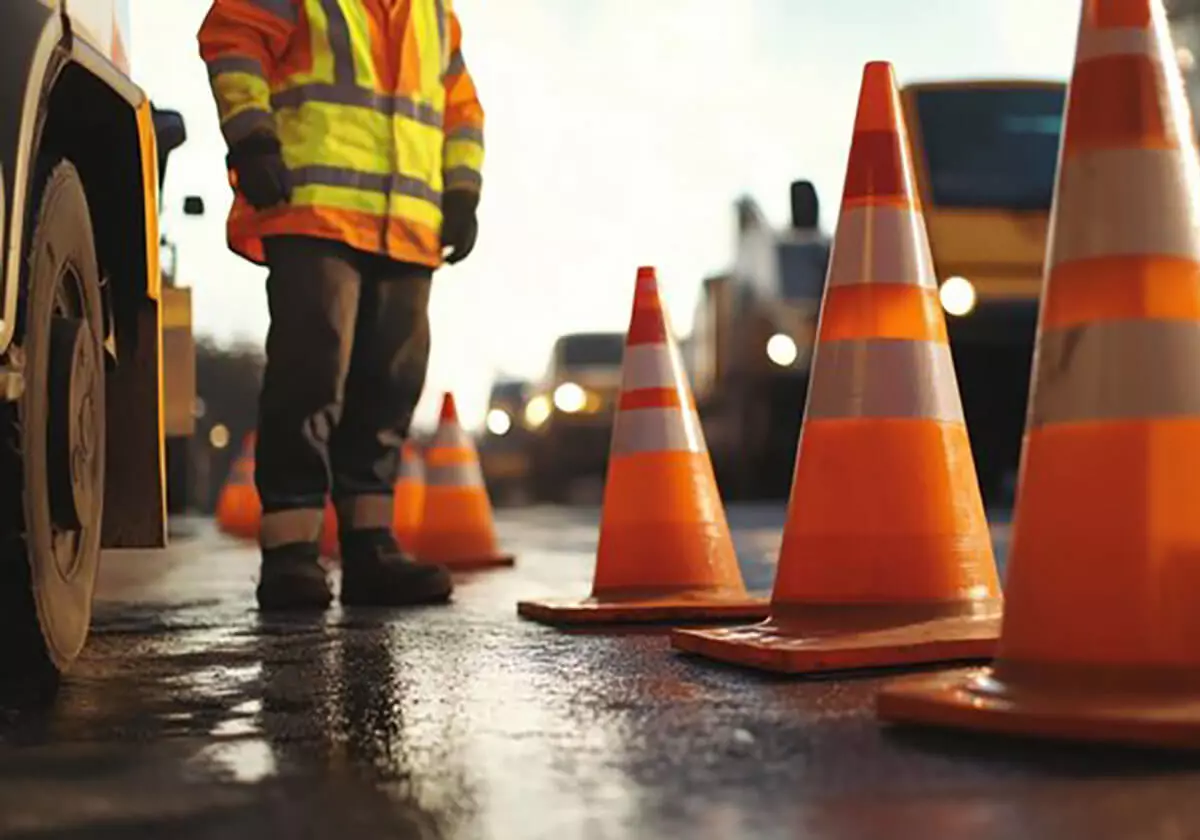 Trabalhador em estrada com cones de sinalização laranja.
